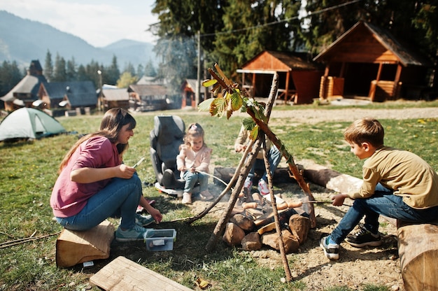 Family bonfire in mountain. frying sausages. mother with four
kids camping. autumn hike and camp weather. warming and cooking
near flame together.