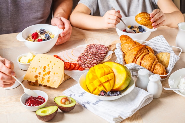 Tavolo per la colazione della famiglia con croissant, marmellata, prosciutto, formaggio, burro, muesli e frutta.