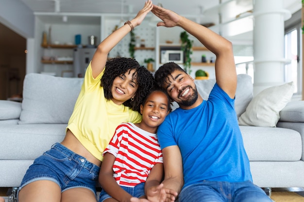 Family Care Protection and Insurance Concept portret van lachende Afro-Amerikaanse ouders die een symbolisch dak van handen maken boven hun gelukkige dochter zittend op het vloertapijt in de woonkamer thuis