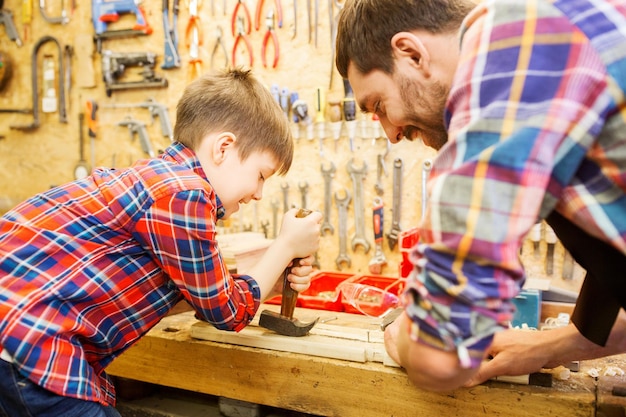 family, carpentry, woodwork and people concept - father and little son with claw hammer pulling nail out of wood plank at workshop