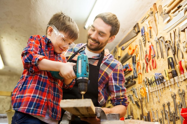 family, carpentry, woodwork and people concept - father and little son with drill perforating wood plank at workshop