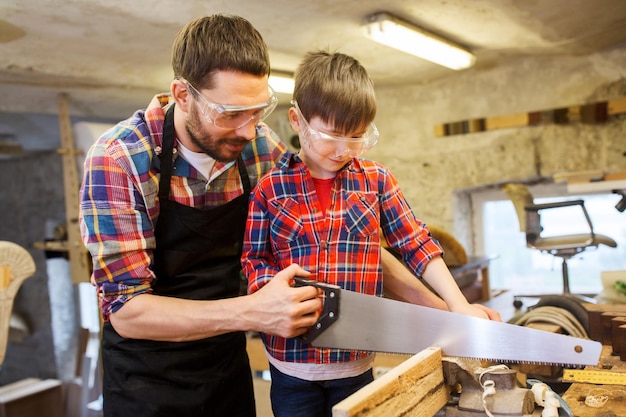 family, carpentry, woodwork and people concept - father and little son with saw sawing wood plank at workshop