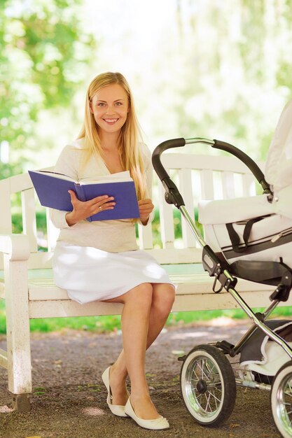 family, child, leisure, education and parenthood concept - happy mother with baby stroller reading book in park