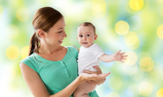 家族と子供と親の概念 - 緑色の休日ライトの背景で小さな赤ちゃんと幸せな笑顔の若い母親