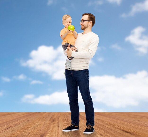 family, childhood, fatherhood, healthy eating and people concept - happy father and and little son with green apple over blue sky and wooden floor background