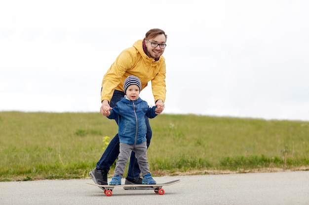 family, childhood, fatherhood, leisure and people concept - happy father teaching little son to ride on skateboard