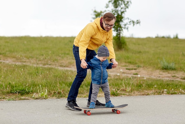 family, childhood, fatherhood, leisure and people concept - happy father teaching little son to ride on skateboard