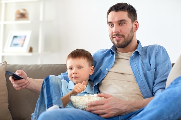 Photo family, childhood, fatherhood, technology and people concept - happy father and little son with remote control and popcorn watching tv at home