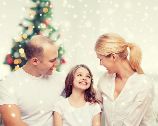 Foto famiglia, infanzia, vacanze e persone - madre, padre e bambina sorridenti sul soggiorno e sullo sfondo dell'albero di natale