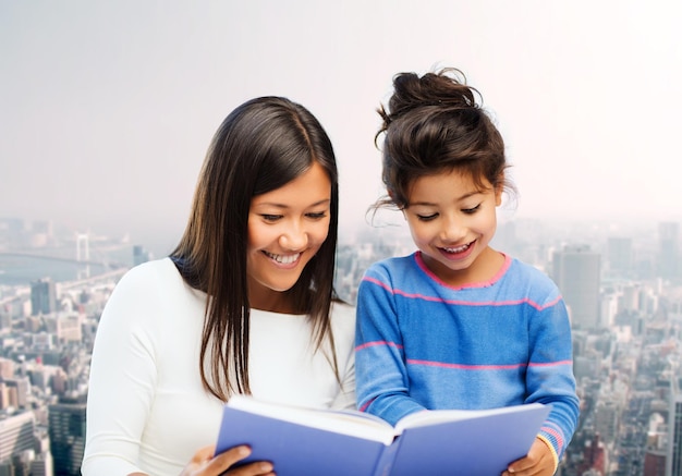 family, children, education and happy people concept - happy mother and little daughter reading book over city background