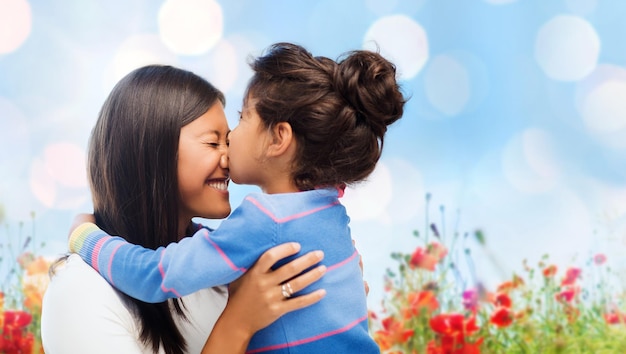 family, children and happy people concept - happy little girl hugging and kissing her mother over blue sky with lights and poppy field background