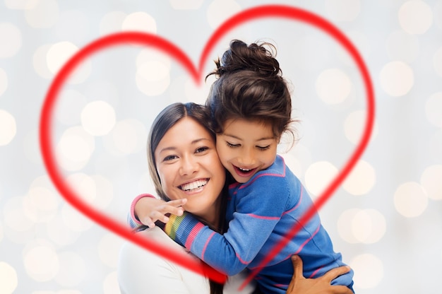 family, children, love and happy people concept - happy mother and daughter hugging over holidays lights background and red heart shape