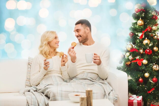 Family, christmas, holidays, love and people concept - happy couple covered with plaid drinking tea and sitting on sofa at home over blue lights background
