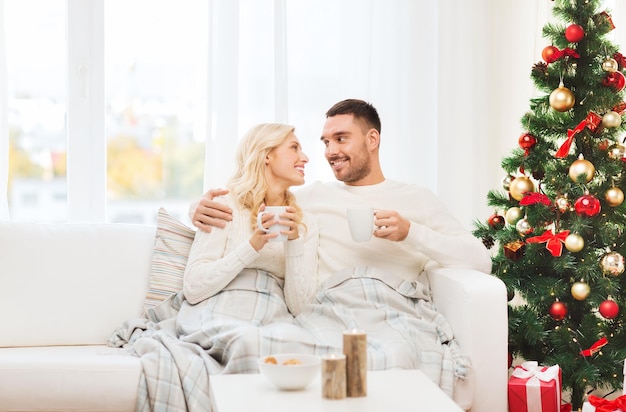 family, christmas, holidays, love and people concept - happy couple covered with plaid drinking tea and sitting on sofa at home