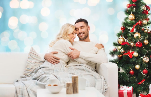 family, christmas, holidays, love and people concept - happy couple covered with plaid sitting on sofa over blue holidays lights background