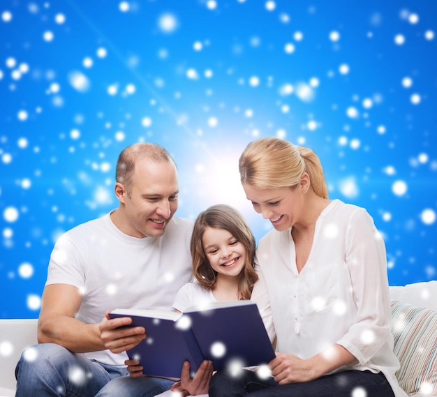 family, christmas, holidays and people - smiling mother, father and little girl reading book over blue snowy background