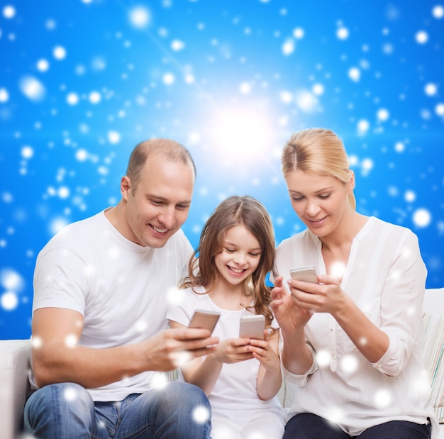 家族、クリスマス休暇、テクノロジー、人々のコンセプト-青い雪の背景の上にスマートフォンで笑顔の母、父、少女