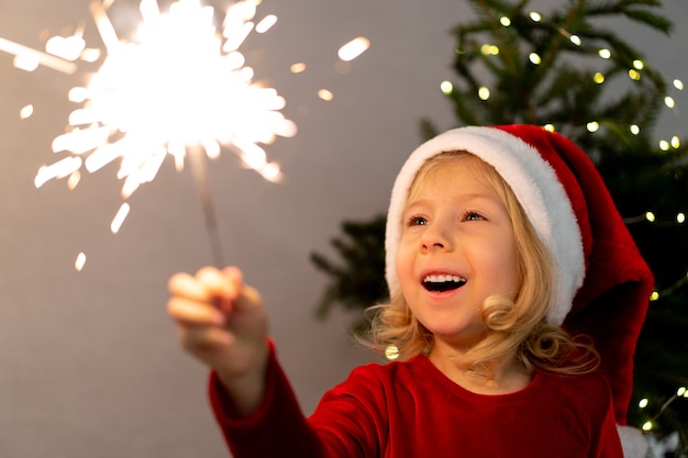 Photo family christmas at home. cute small girl in santa hat holding burning sparkler and smile happily over defocused christmas background, indoor emotional lifestyle.