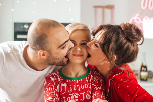 Family in Christmas pajamas in the kitchen cheerfully bite the boy's cheeks