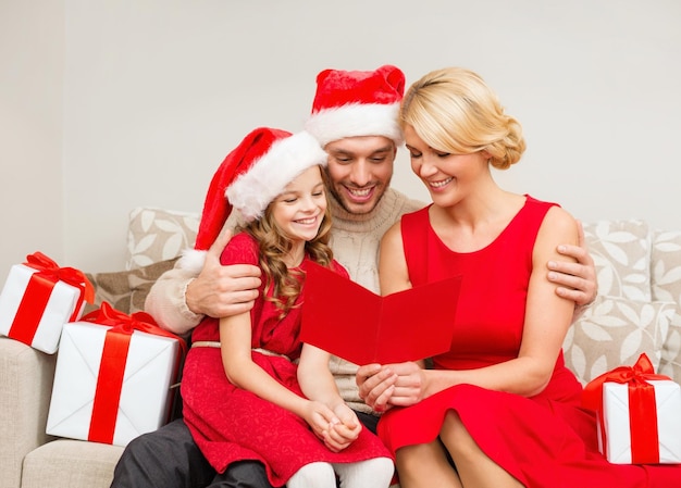 family, christmas, x-mas, happiness and people concept - smiling family in santa helper hats with many gift boxes reading postcard