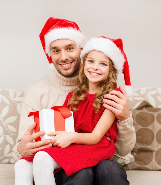 family, christmas, x-mas, winter, happiness and people concept - smiling father and daughter in santa helper hats holding gift box