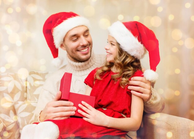 家族、クリスマス、クリスマス、冬、幸福と人々のコンセプト-サンタヘルパー帽子の笑顔の父と娘がギフトボックスを開く