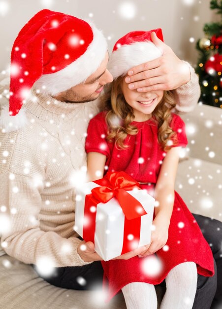 family, christmas, x-mas, winter, happiness and people concept - smiling father surprise daughter with gift box covering eyes with hand