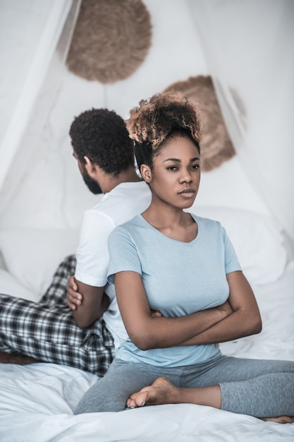 Семья, конфликт. Сердитая афро-американская женщина со скрещенными руками на груди сидит спиной к мужу в спальне на кровати