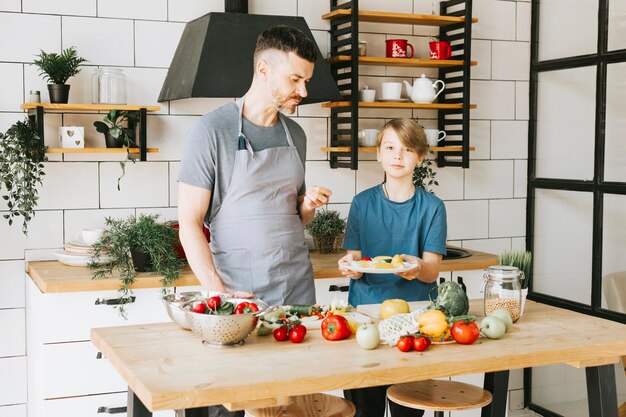 写真 家族の父親の若者と息子の10代の少年がキッチンで野菜サラダを作り、一緒に充実した時間を過ごす父と息子が話したり、ベジタリアン料理を作ったり、家事をしたりする3月8日と母の日