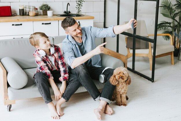 家族のお父さんの若者と息子の十代の少年は、ペットのプードル犬と一緒に居心地の良いアパートのソファに座り、電話で自分撮りをして家族と充実した時間を過ごしています