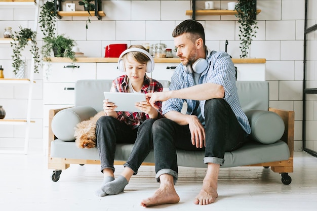 family dad young man with phone and son teenage cute boy with tablet and headphones sit on couch in cosy apartment with pet poodle dog quality time with your family gadget addicted