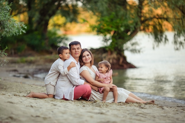 家族の日、幸せな親。ママ、パパ、息子、娘が散歩を楽しんでいます。彼らは川沿いの砂の上に座っています