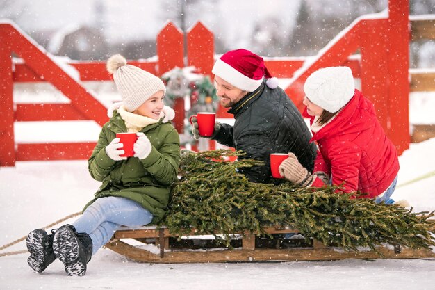 Family drinking tea to keep warm on a sleigh with a Christmas tree in a snowy winter outside