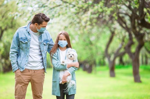 Family of father and little daughter with puppy wearing protective medical mask outdoors in the park