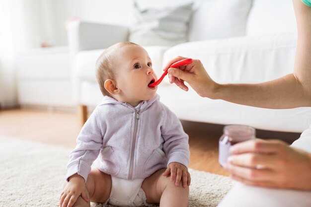 家族、食べ物、子供、食事、親子関係のコンセプト – 家庭で小さな赤ちゃんに餌を与えるピューレとスプーンを持つ母親