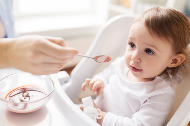 家族、食べ物、食事、人々のコンセプト – 自宅のハイチェアに座っている小さな赤ちゃんにピューレとスプーンで餌を与える母親