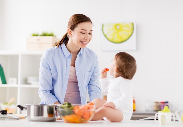 家族、食べ物、健康的な食事、料理、人々のコンセプト – 家庭のキッチンでニンジンを持つ幸せな母親と小さな女の赤ちゃん