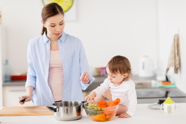 family, food, healthy eating, cooking and people concept - happy mother and little baby girl with vegetables and pot at home kitchen