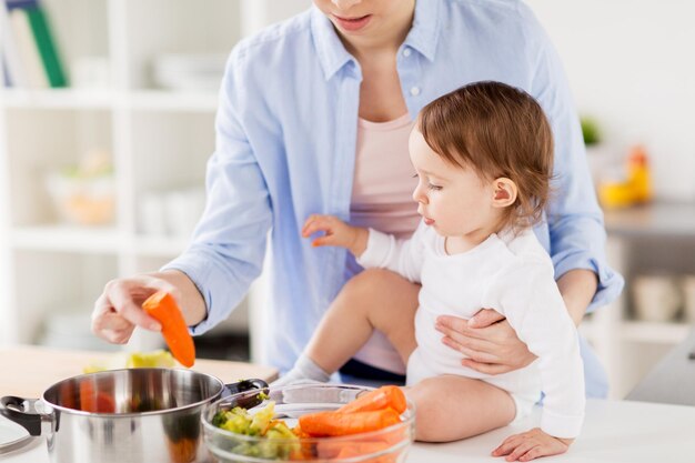 家族食事健康的な食事調理人とのコンセプト - 家のキッチンで野菜と鍋を持った幸せな母親と小さな赤ちゃん