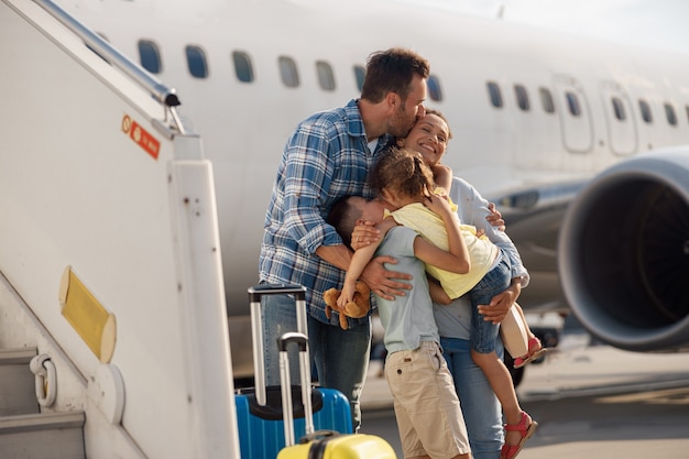 Семья из четырех человек целует друг друга во время поездки, стоя перед большим самолетом на открытом воздухе. Люди, путешествия, концепция отпуска