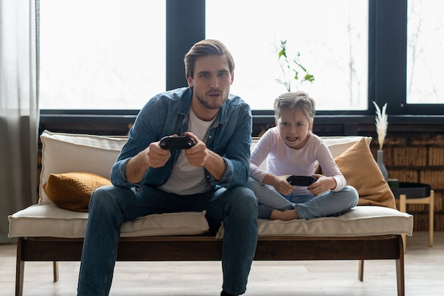 Concetto di famiglia, gioco e intrattenimento: padre felice e figlia piccola con gamepad che giocano a videogiochi a casa.