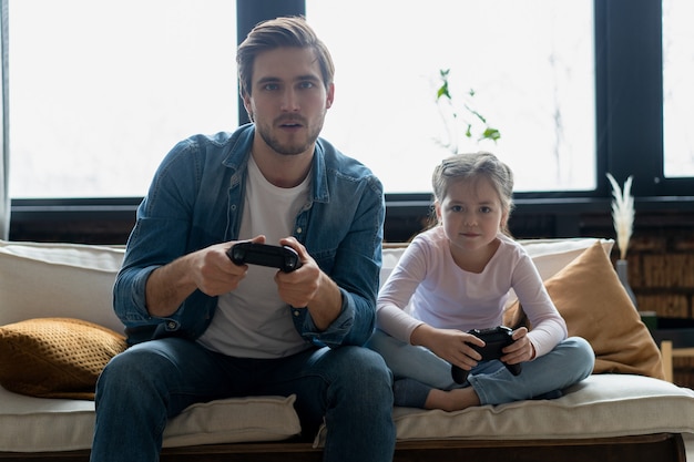 Family, gaming and entertainment concept - happy father and little daughter with gamepads playing video game at home.