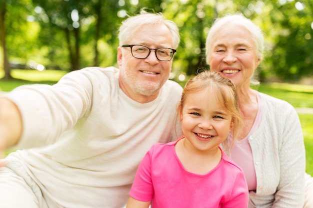 family, generation and people concept - happy smiling grandmother, grandfather and little granddaughter taking selfie at park