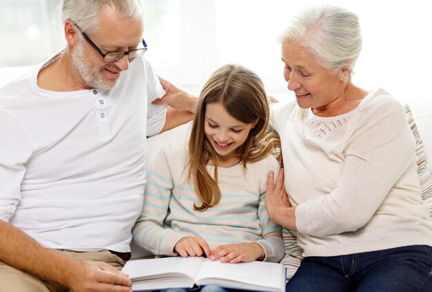 family, generation and people concept - smiling grandfather, granddaughter and grandmother with book sitting on couch at home