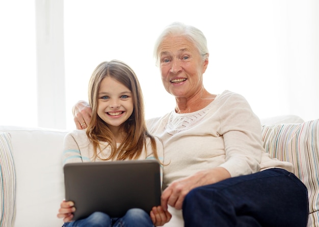концепция семьи, поколения, технологий и людей - улыбающиеся внучка и бабушка с планшетным компьютером сидят дома на диване