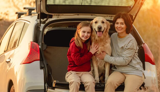 Family girl and child with golden retriever dog sitting in car\
trunk outdoors. young mother and daughter kid with doggy pet\
labrador in vehicle at nature at autumn