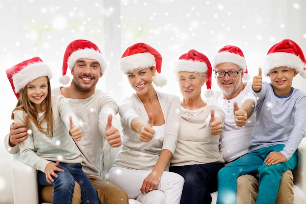 family, happiness, generation, holidays and people concept - happy family in santa helper hats sitting on couch and showing thumbs up gesture at home