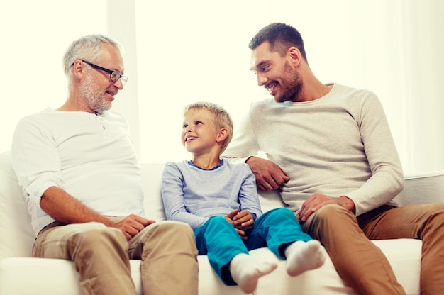 family, happiness, generation and people concept - smiling father, son and grandfather sitting on couch at home