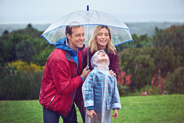 Photo family happy and outdoor in rain with umbrella in nature for fun happiness or quality time man woman and excited child with cover for water drops with freedom love and care while playing at park