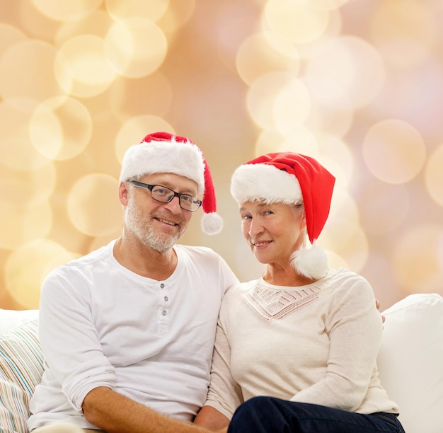 family, holidays, christmas, age and people concept - happy senior couple in santa helper hats sitting on sofa over beige lights background